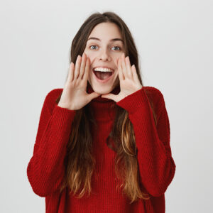 Excited young female with long dark hair exclaiming, looking at camera with happy expression, holding hands behind opened mouth, calling someone, being glad. Positive emotions, body language.