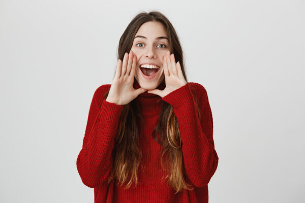 Excited young female with long dark hair exclaiming, looking at camera with happy expression, holding hands behind opened mouth, calling someone, being glad. Positive emotions, body language.