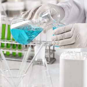 Young clinical worker dressed in white gown and gloves holding glass beakers with blue liquid making experiment in laboratory. Scientific laboratory research, laboratory beakers in hadns of scientist